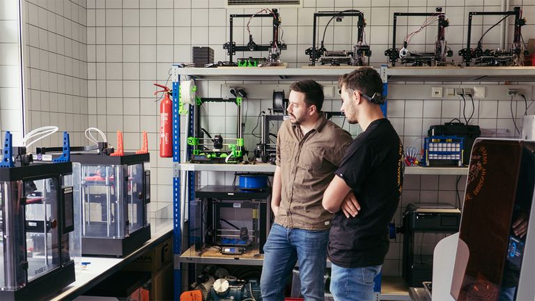 3D-Drucker in einem Regal, zwei Personen stehen davor