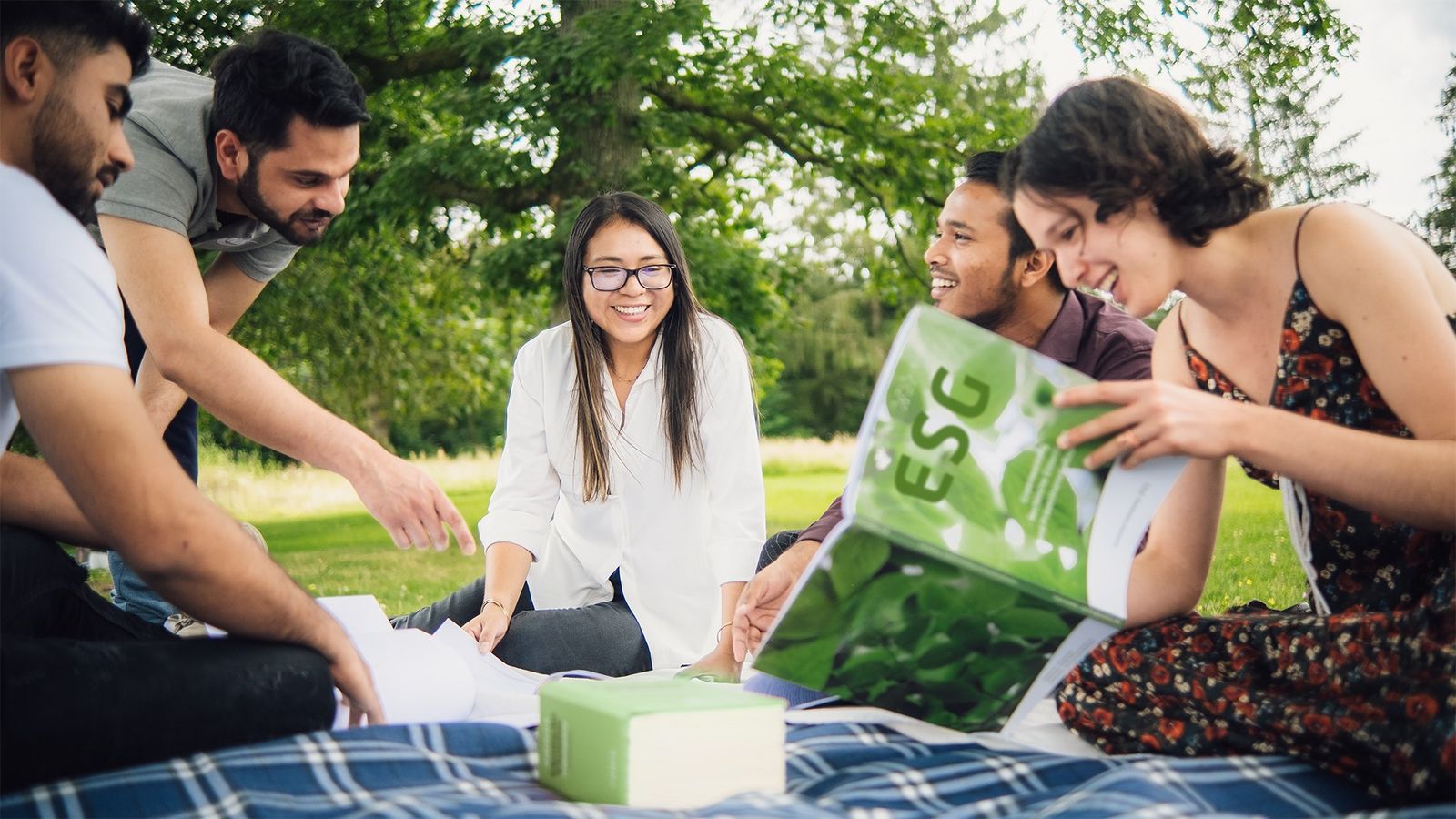 Gruppe internationaler Studierender sitzt im Freien auf einer Picknick-Decke und diskutiert über ein ESG-Buch.